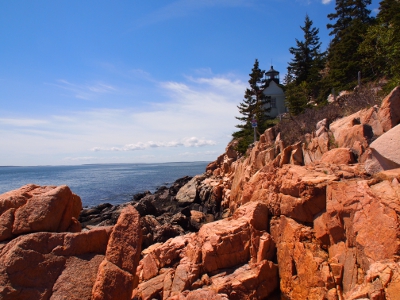 [Salmon colored rock in foreground with dark rock in center and evergreens and a lighthouse on the hillside on the right. The ocean and clouds in the sky are seen in the distance.]